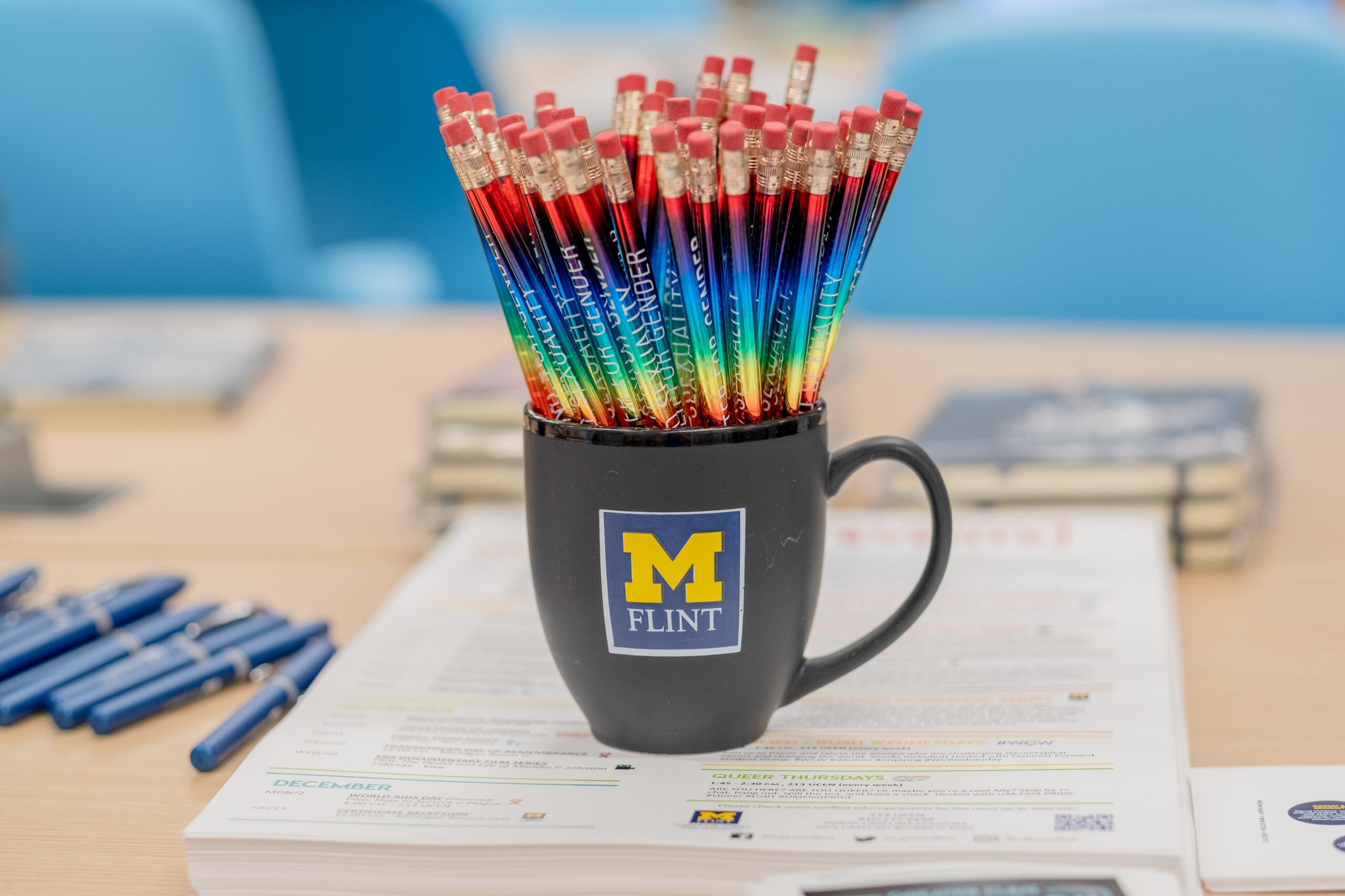 A black mug with "M FLINT" logo filled with colorful pencils, placed on a table with papers and pens nearby.