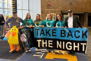 A group of five people holds a large banner reading "TAKE BACK THE NIGHT," with one person in a duck costume nearby.