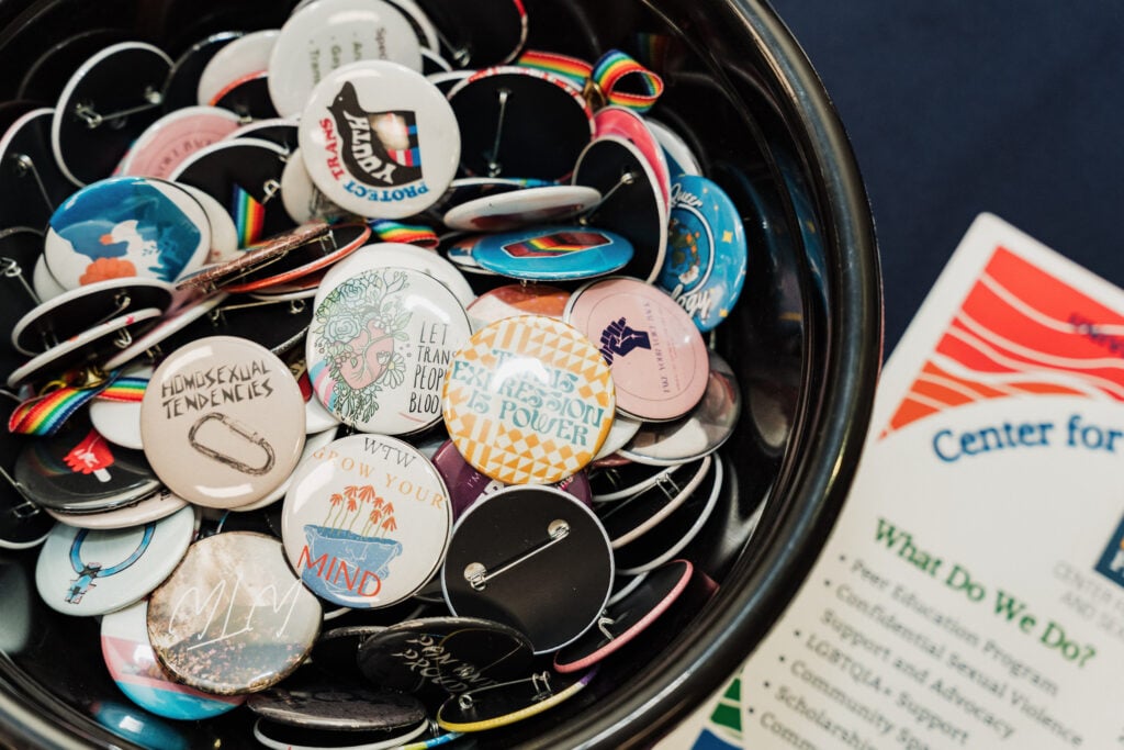 A bowl filled with various colorful buttons featuring messages and designs related to LGBTQ+ themes, alongside a pamphlet.