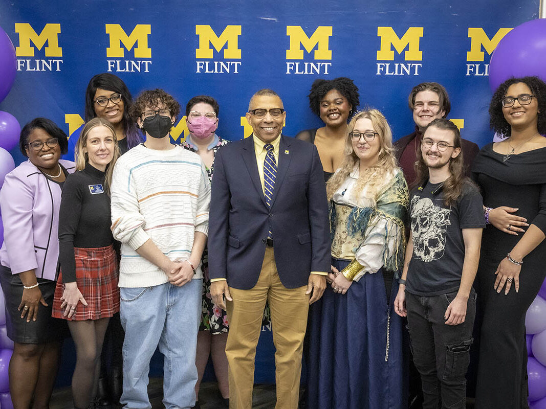 A group of ten people poses together in front of a blue backdrop with "M FLINT" and purple balloons, smiling for the camera.