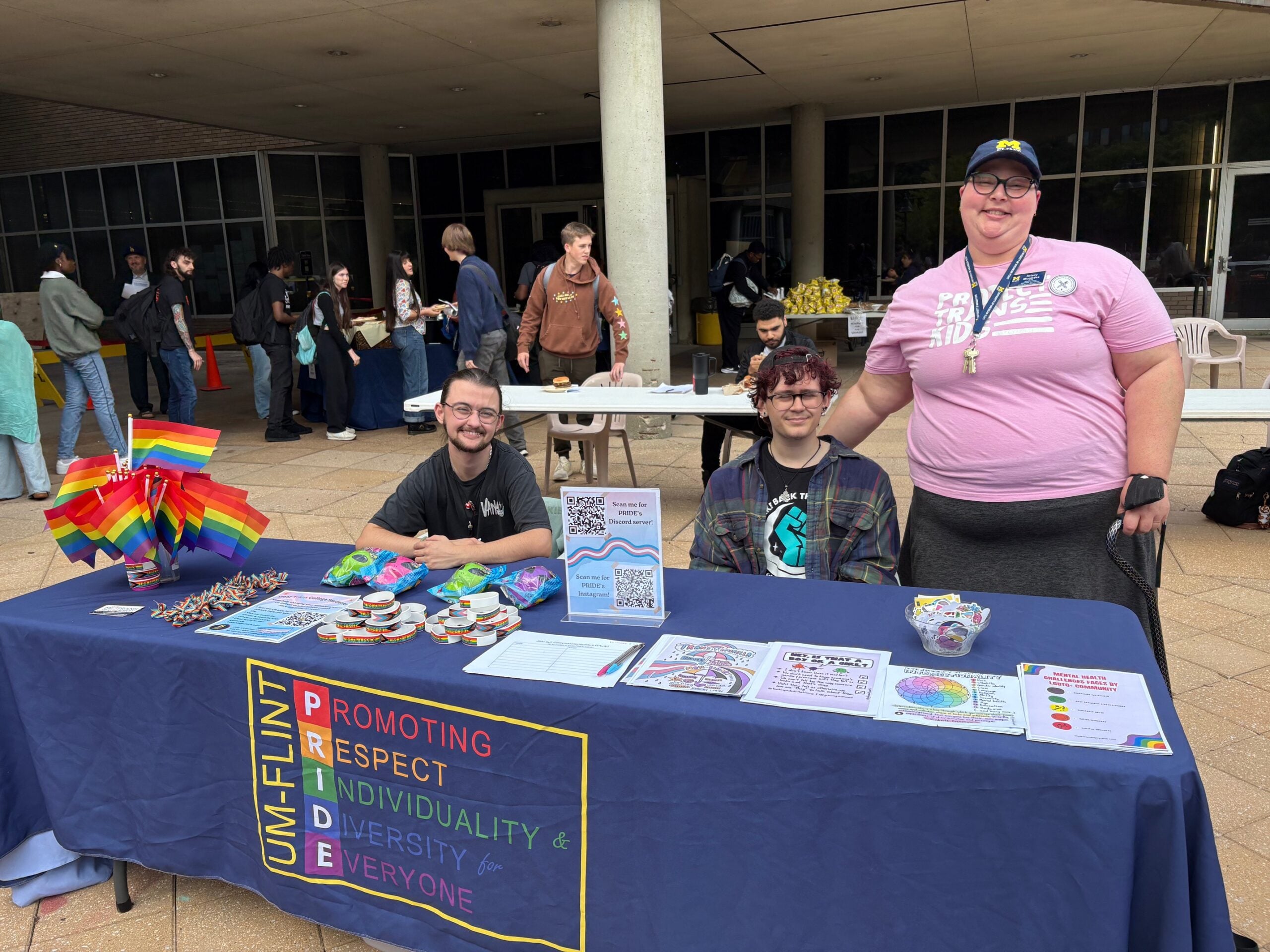 Three people are seated at a table with a rainbow flag and various informational materials promoting diversity and pride.