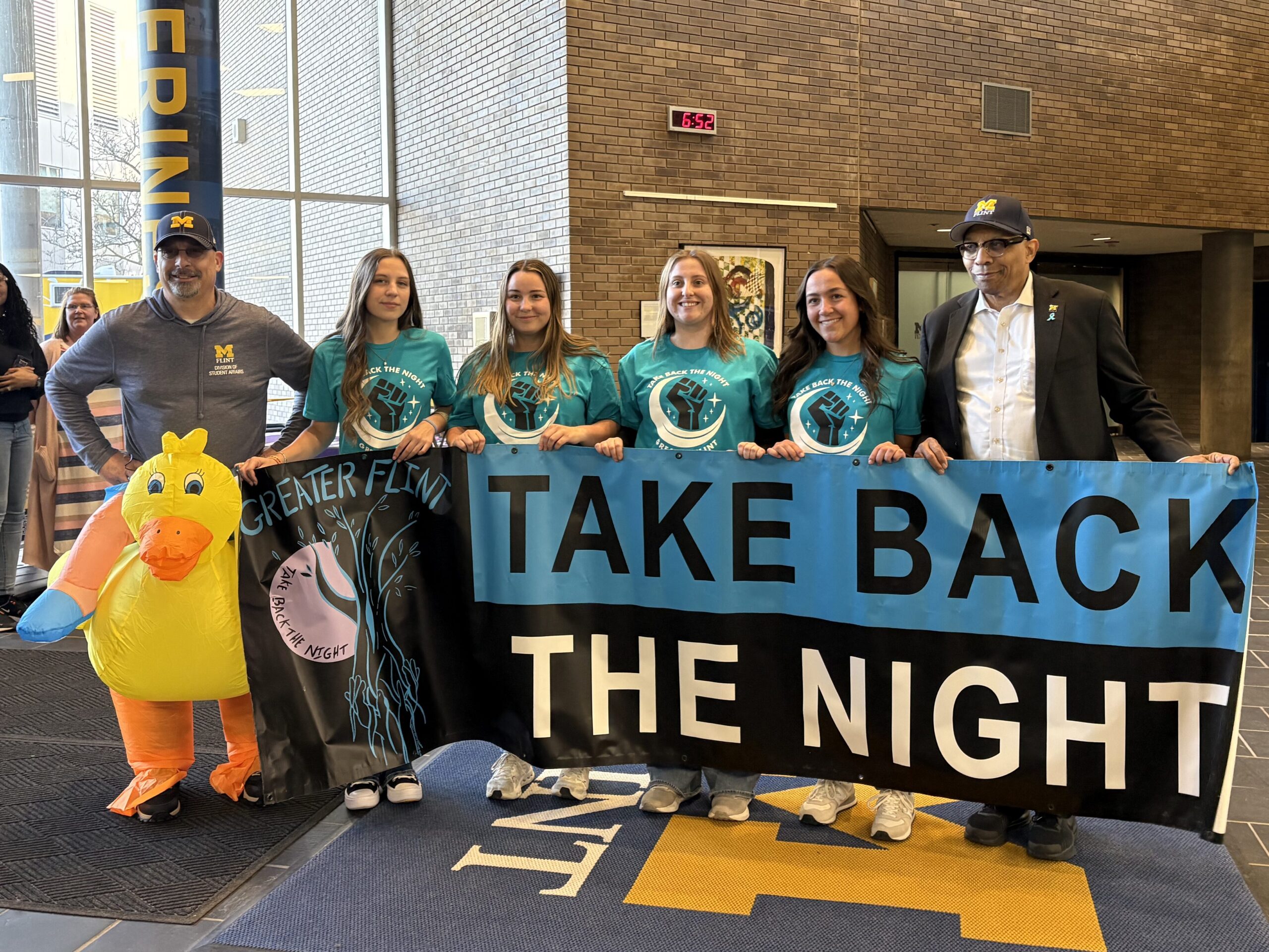 A group of people stands in a lobby holding a banner that reads "TAKE BACK THE NIGHT," with a person in a duck costume nearby.