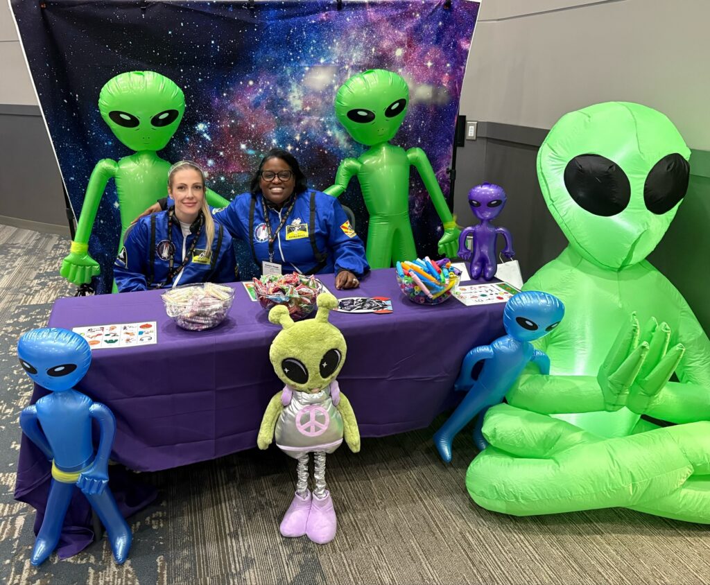 Two people sit at a table decorated with alien-themed items, surrounded by inflatable alien figures and a colorful display.