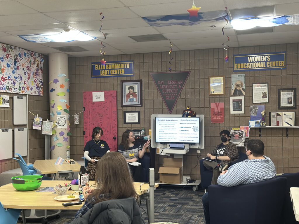 A group of people sits in a colorful room with educational posters, discussing topics displayed on a screen.