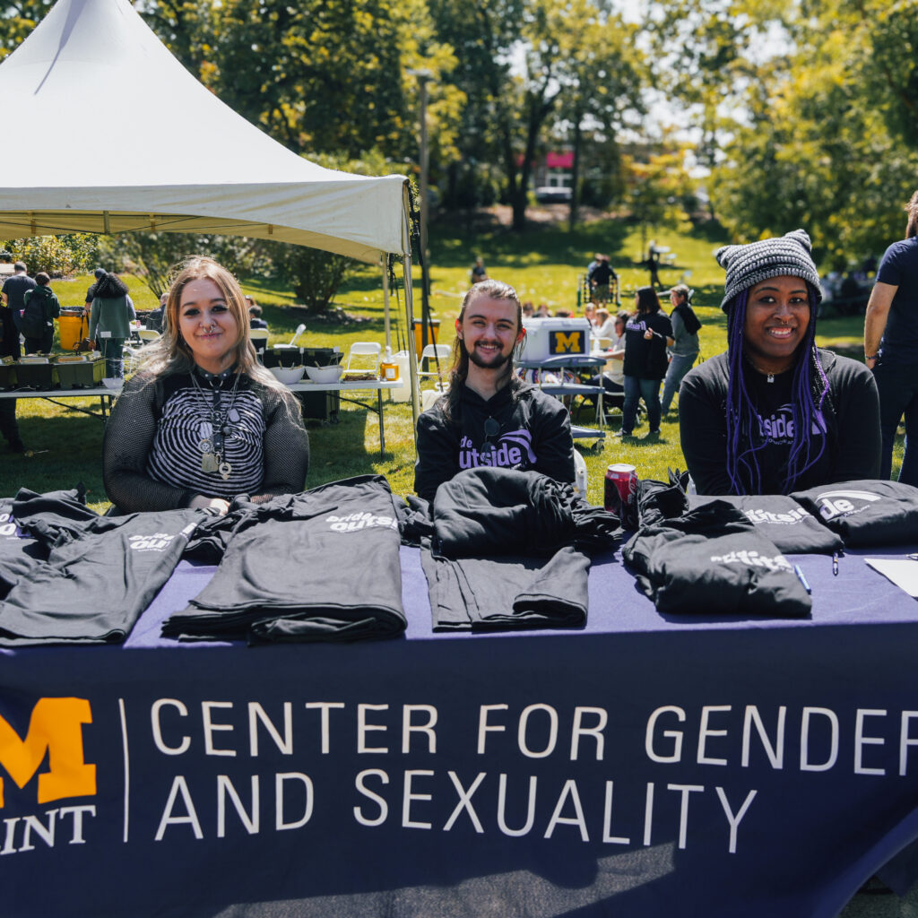 Three people sit behind a table displaying T-shirts, promoting the Center for Gender and Sexuality at an outdoor event.