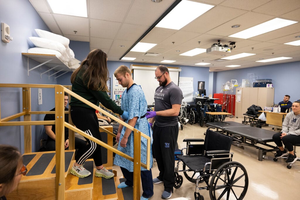 Physical therapy students practice helping a patient in a hospital gown climb stairs while classmates observe in a rehabilitation lab.