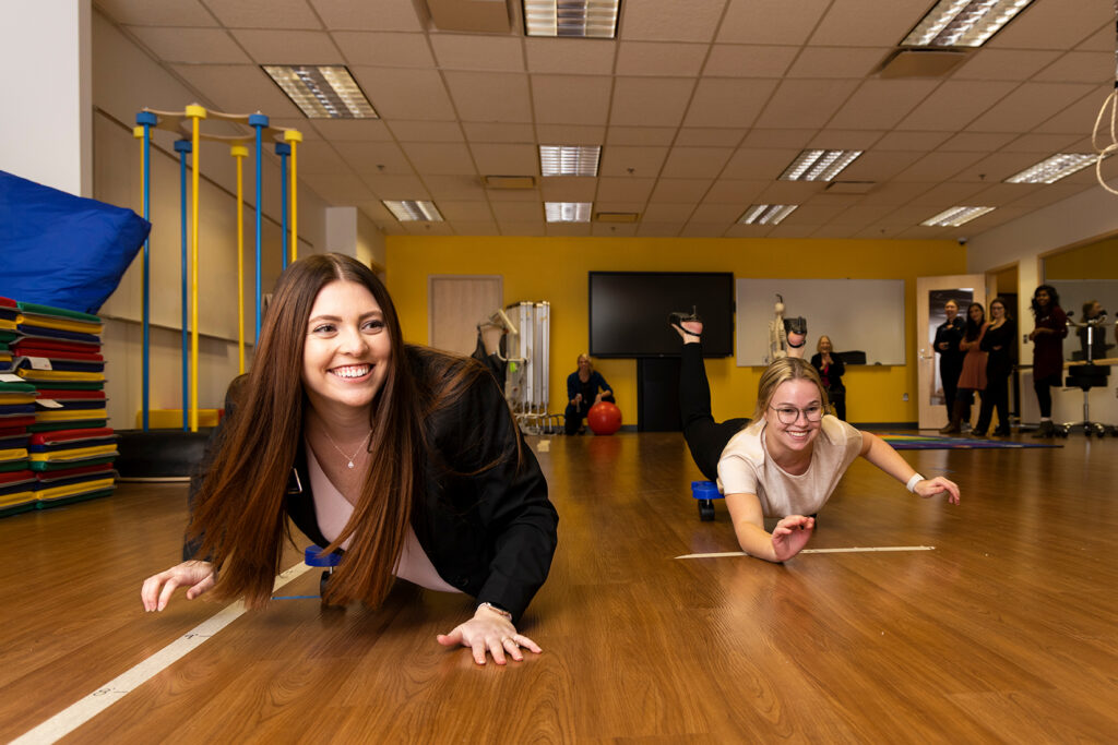 Two UM-Flint students smile as they propel themselves across a laboratory floor on wheeled scooter boards while faculty members watch.