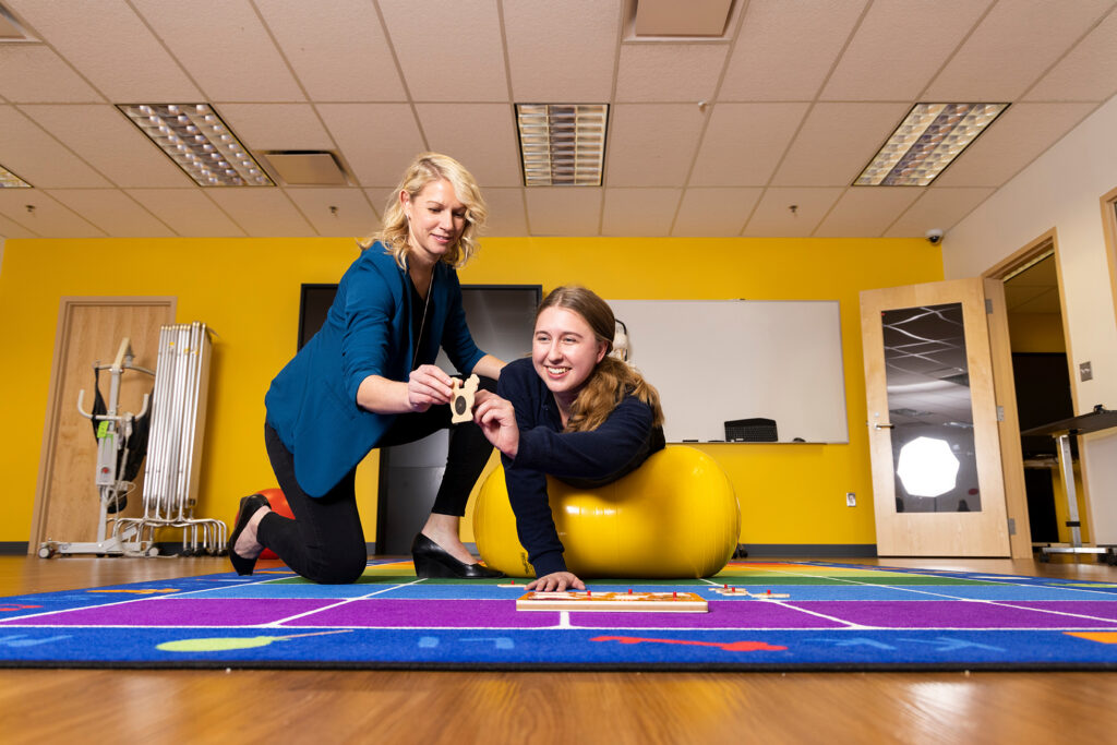 A UM-Flint occupational therapy student balances over an exercise ball while placing a wooden puzzle piece with guidance from an instructor.