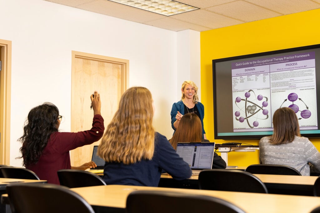 An occupational therapy instructor leads a classroom discussion as a student raises her hand and others follow along on laptops.
