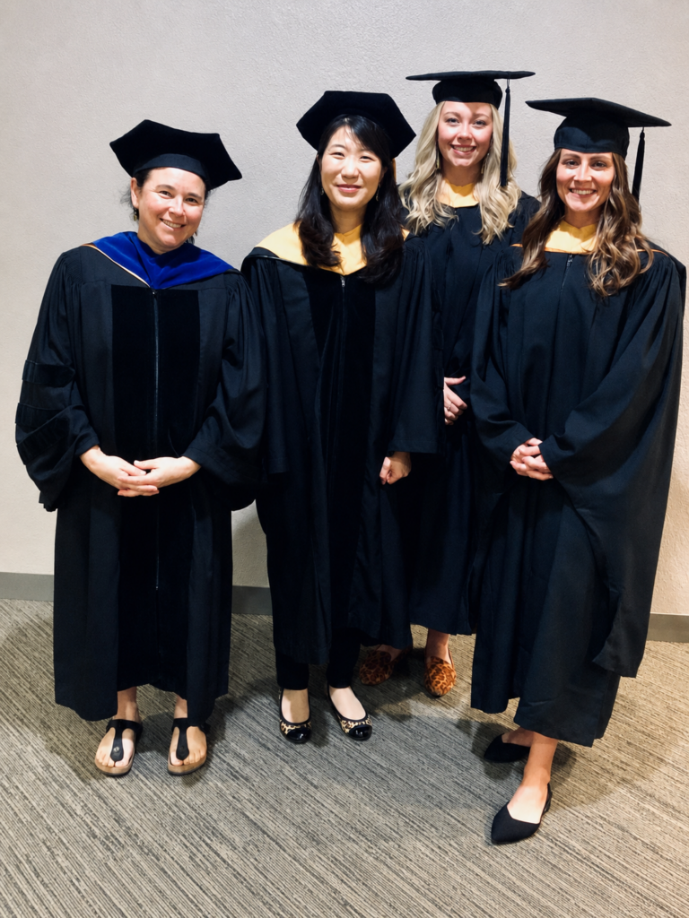 Four individuals in graduation gowns and caps stand together, smiling in a formal setting.