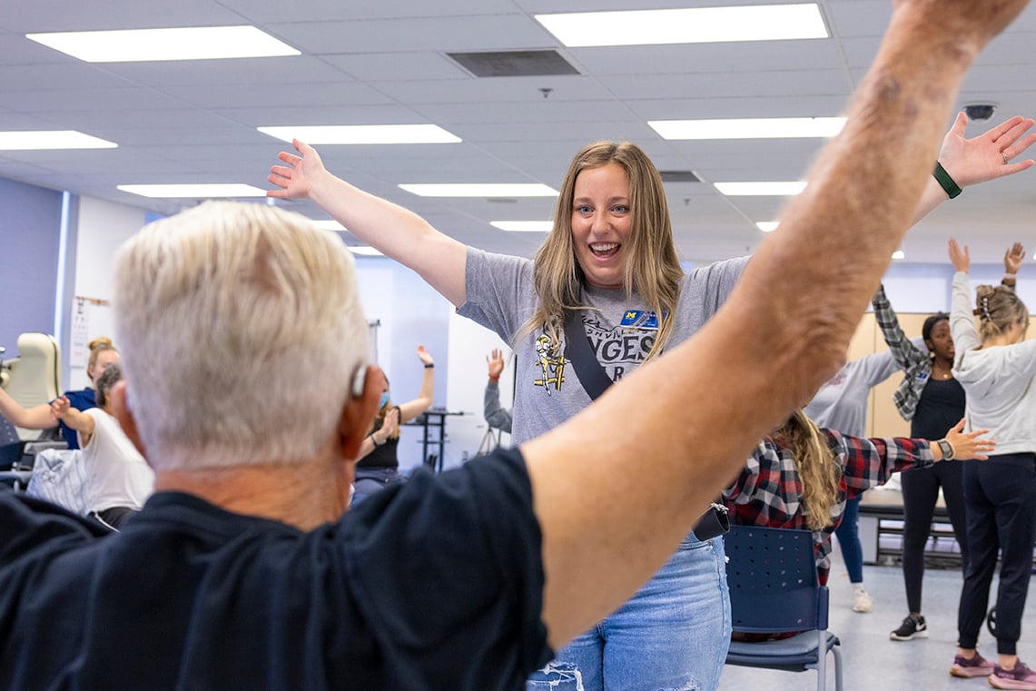 A student raises her arms while participating in a seated movement activity with an older adult and classmates in a university classroom.