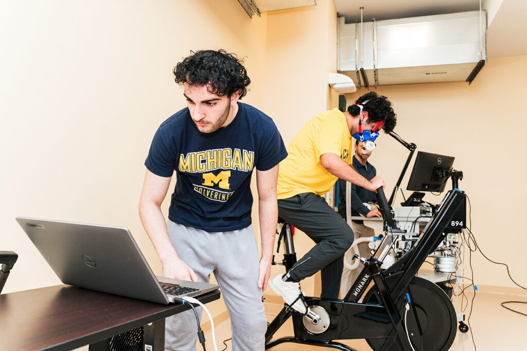 An exercise science student wearing a respiratory testing mask pedals a stationary bike while another student and an instructor monitor the assessment.
