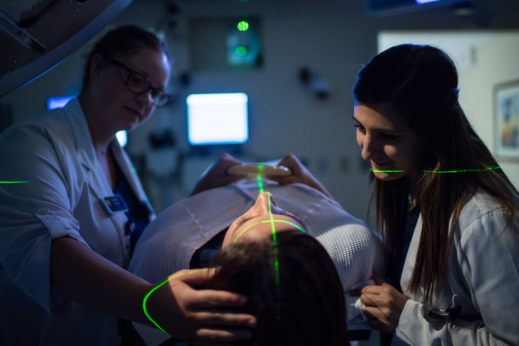 Two UM-Flint radiation therapy students position a patient beneath treatment equipment as green alignment lasers cross the patient’s face and body.