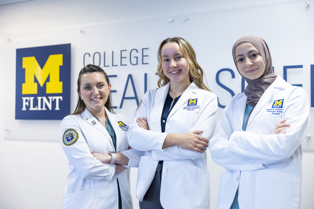 Three UM-Flint physician assistant students in white coats pose with arms crossed in front of the College of Health Sciences sign.