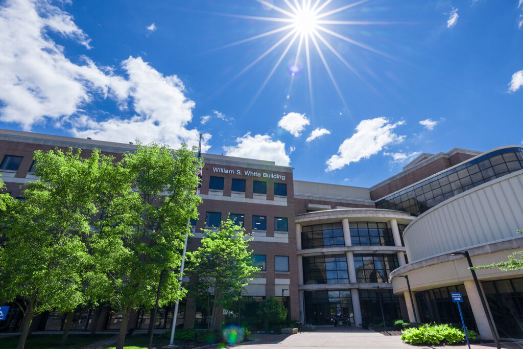 The William S. White Building at UM-Flint stands beneath a bright blue sky with sunlight, clouds and leafy trees.