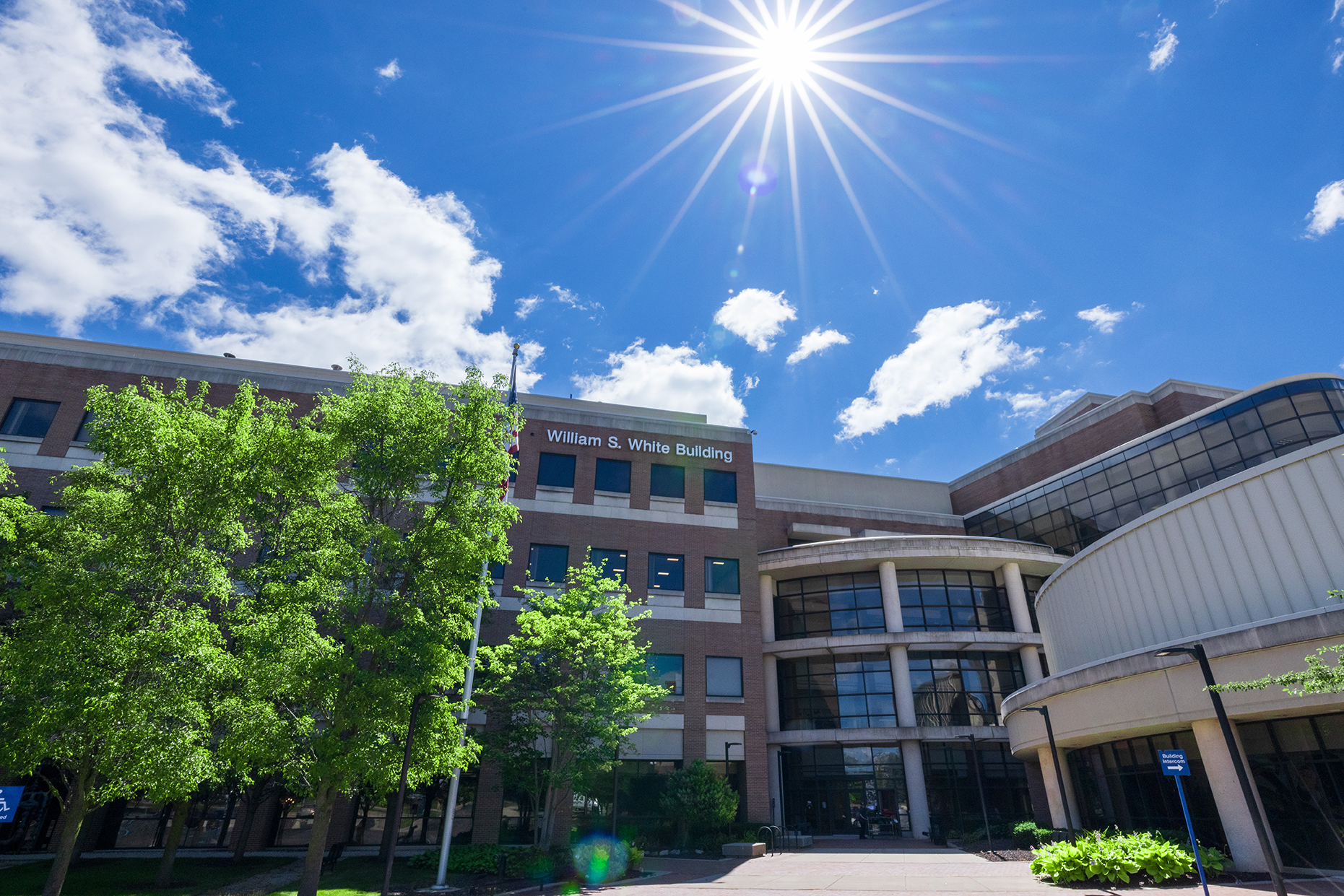 The William S. White Building at UM-Flint stands beneath a bright blue sky with sunlight, clouds and leafy trees.