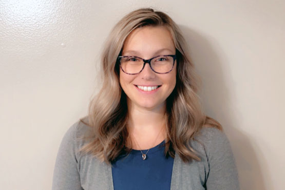 A woman with glasses smiles for a headshot, wearing a blue top and gray cardigan against a neutral background.