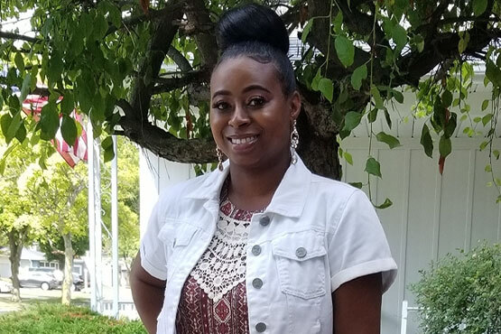 A woman smiles outdoors beneath a leafy tree, wearing a white denim jacket over a patterned top.
