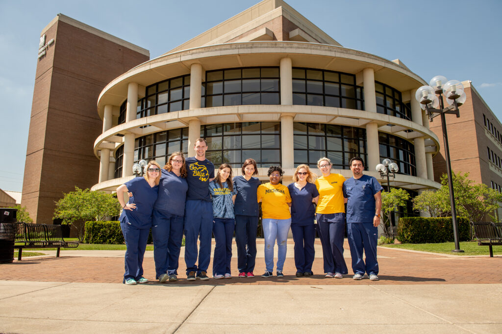 Nine UM-Flint radiation therapy students and faculty members pose together outside a campus building, wearing navy and maize apparel.