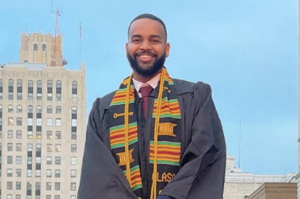 A UM-Flint graduate smiles in a black cap and gown with a colorful Kente stole and gold honor cords, with downtown Flint buildings in the background.