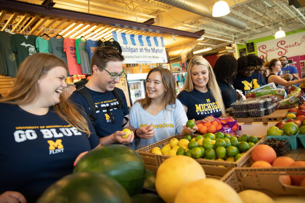 A group of four people smiles and interacts while selecting fruits at a market, with colorful produce displayed in baskets.