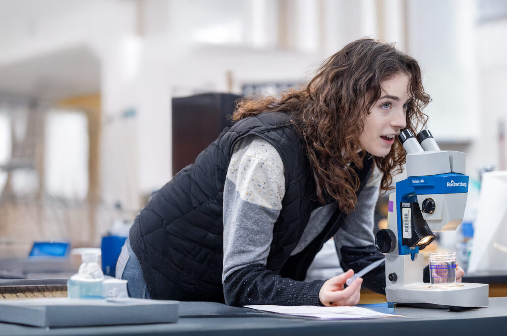 A young female college student looks up from her microscope and smiles while conducting research at the College of Innovation and Technology.