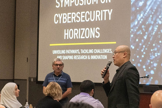 A speaker addresses an audience at the Symposium on Cybersecurity Horizons event, with a presentation slide in the background.