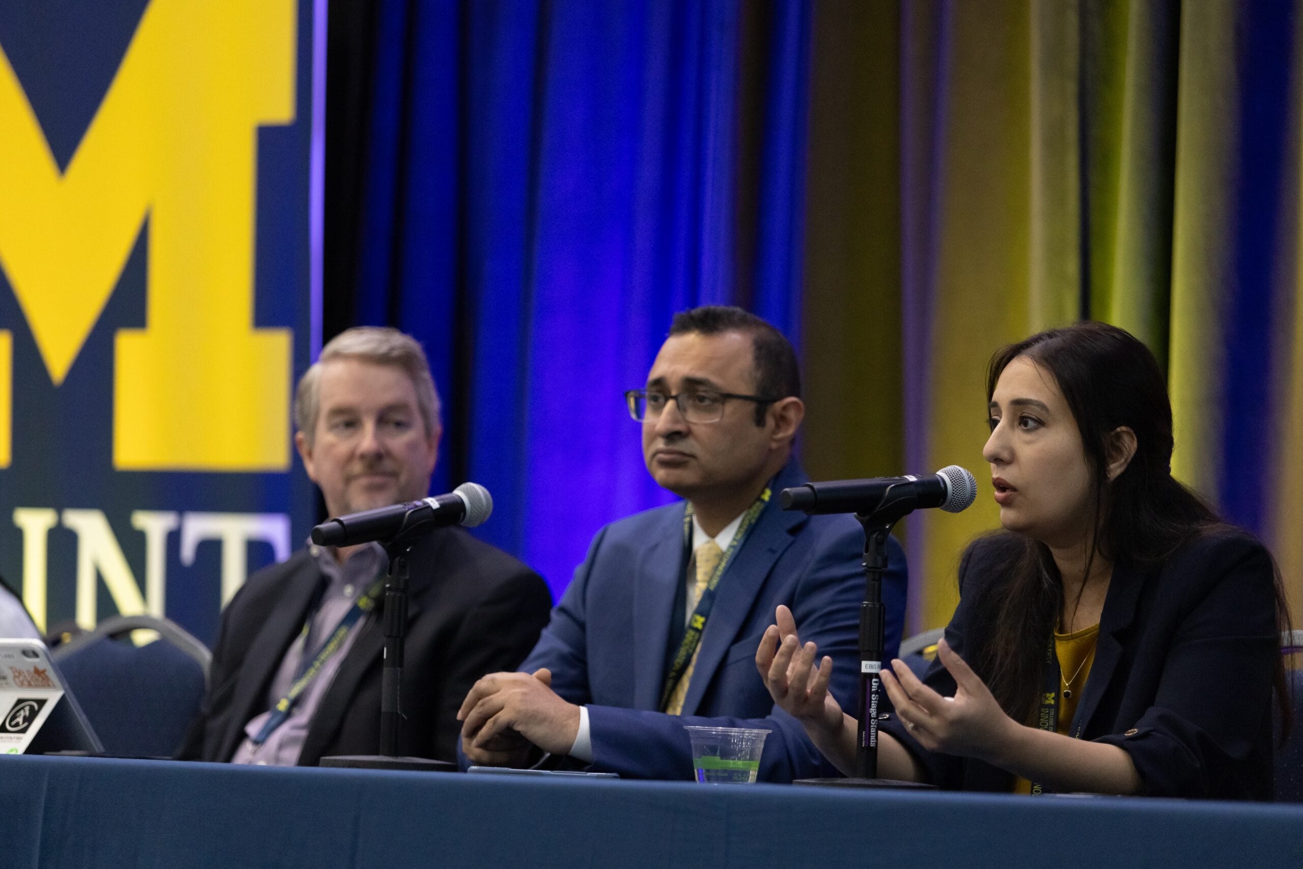 Three people sit at a panel discussion, with microphones in front of them, discussing a topic related to the event.
