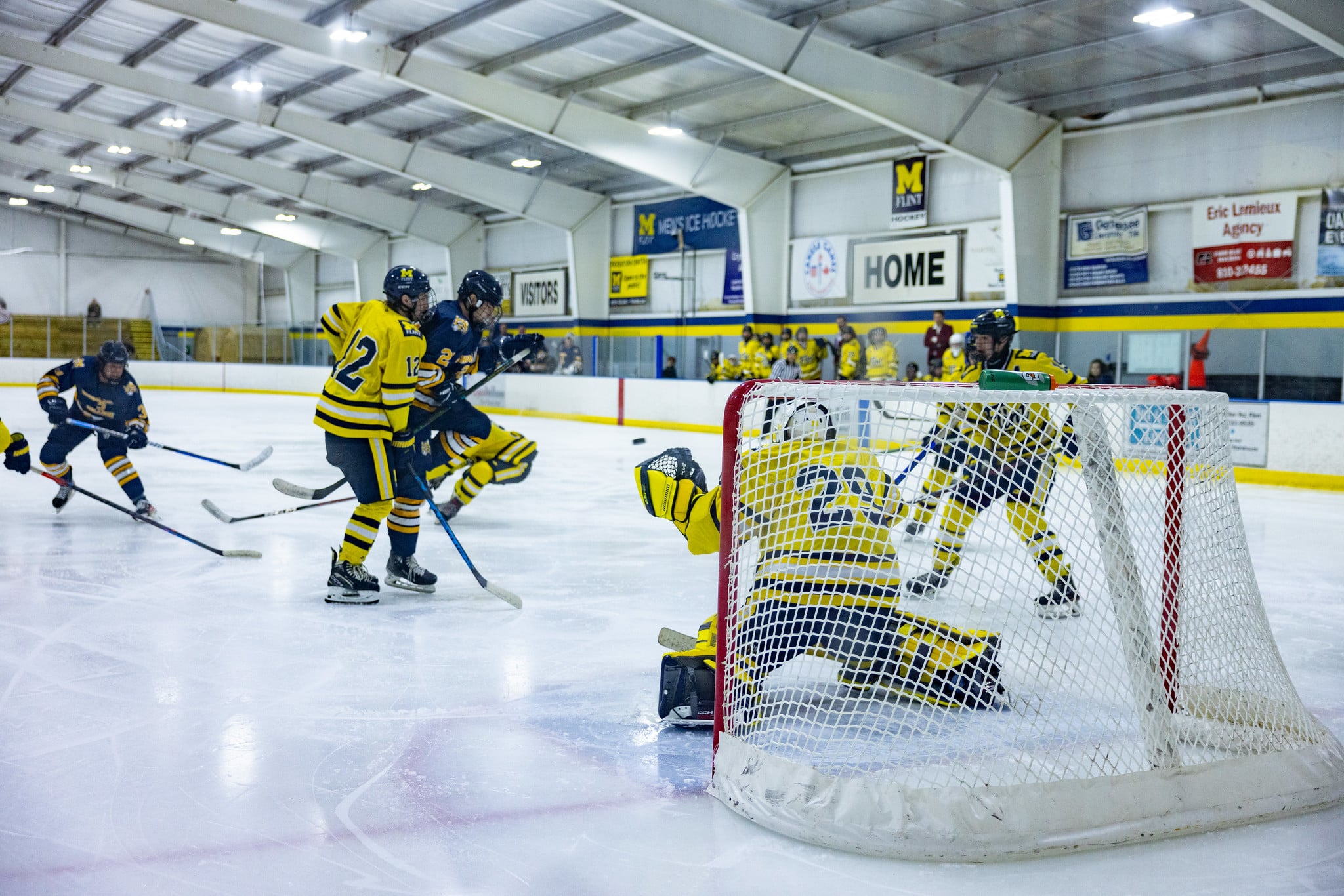 A hockey game in progress with players in yellow and blue jerseys, one player attempting a shot on goal while the goalie defends.