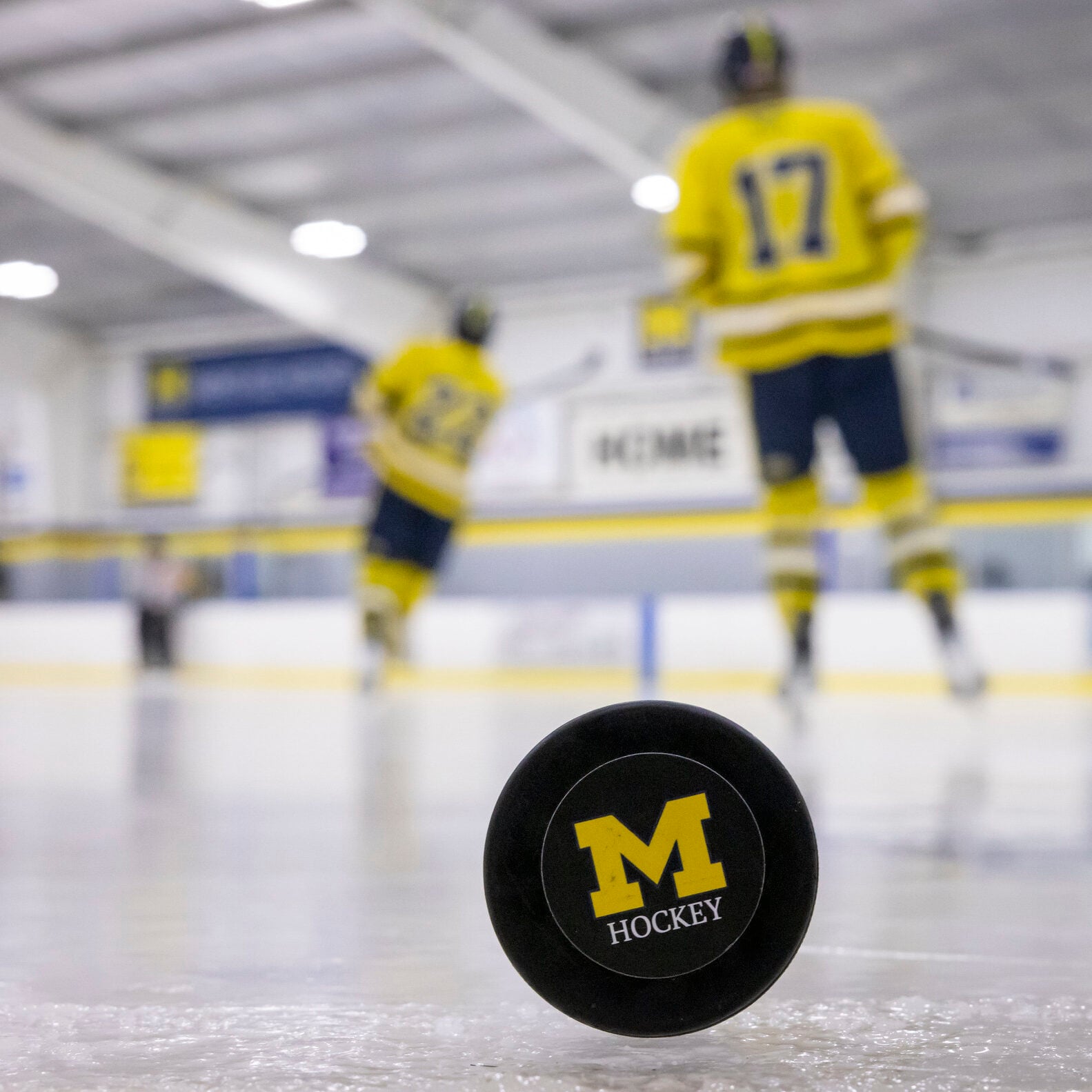 A hockey puck with a yellow "M HOCKEY" logo in the foreground, with blurred players in yellow jerseys skating in the background.