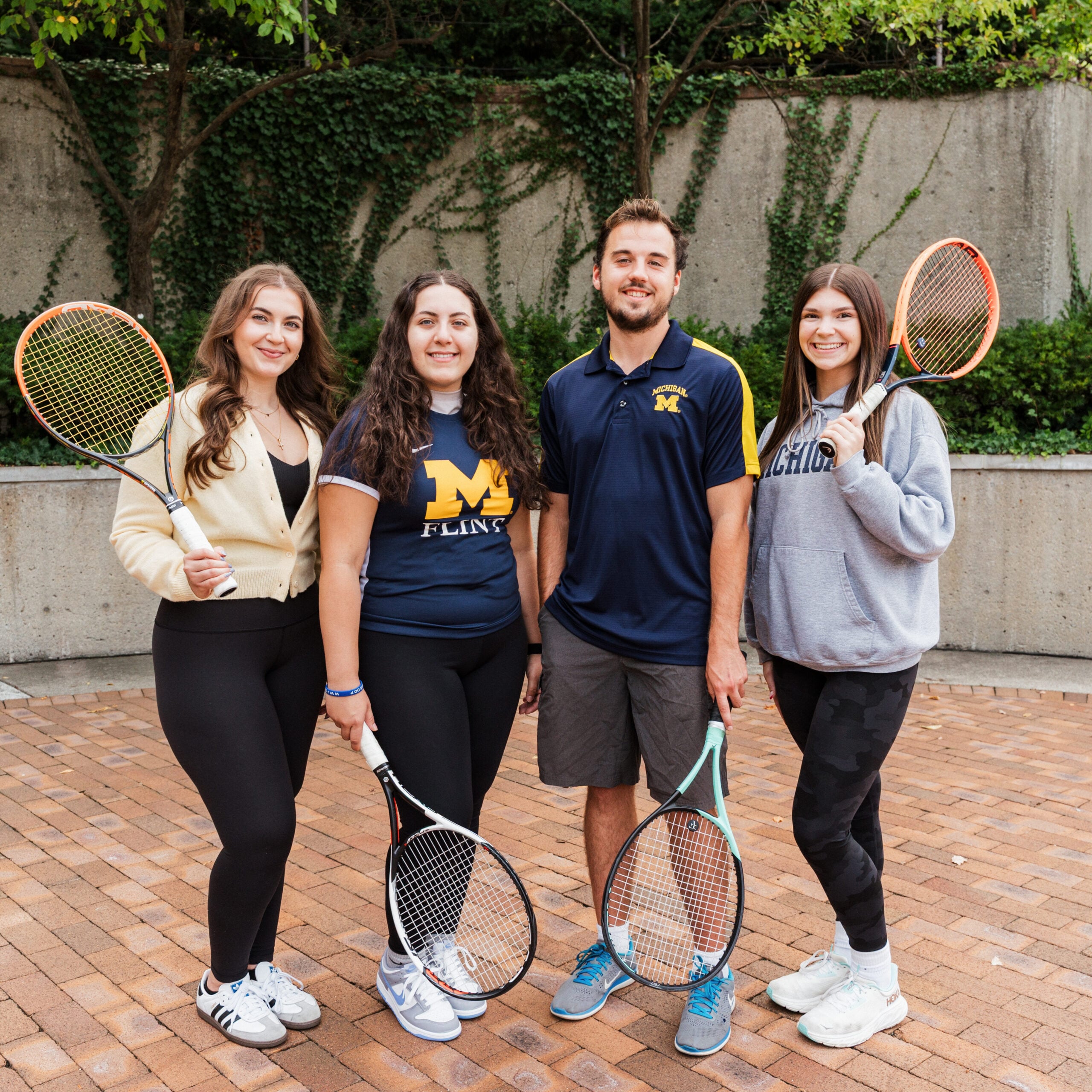 Four people stand together outdoors, each holding a tennis racket, with greenery and a stone wall in the background.
