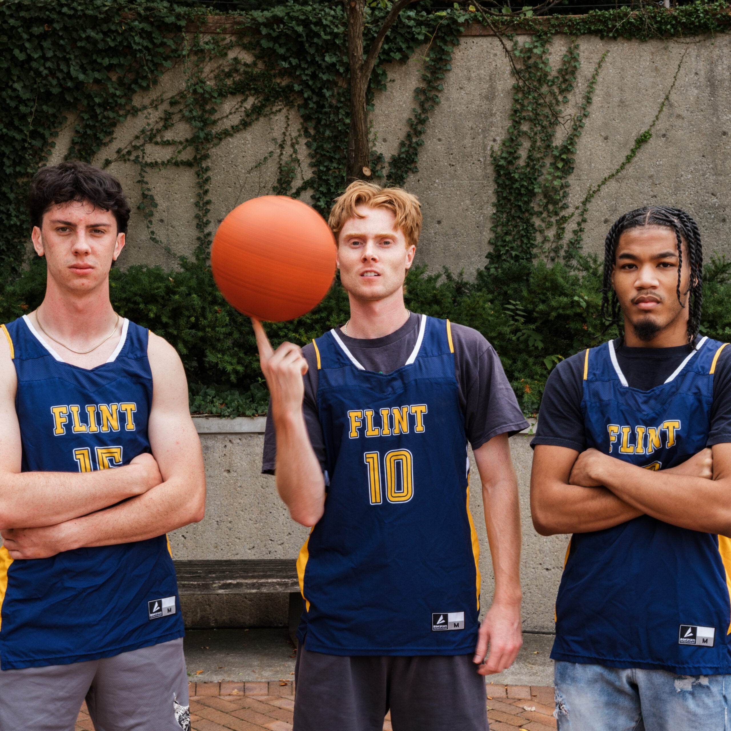 Three people in basketball jerseys stand together outdoors, with one person spinning a basketball on their finger.