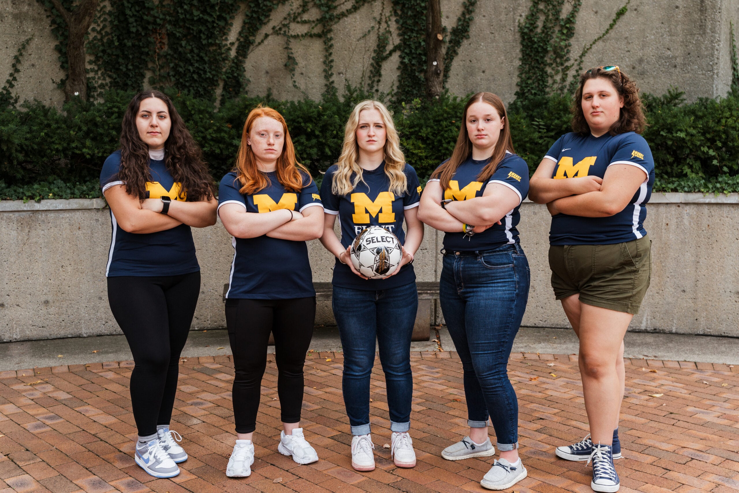 Five people stand confidently in a row, wearing matching blue shirts with a yellow "M," holding a soccer ball.
