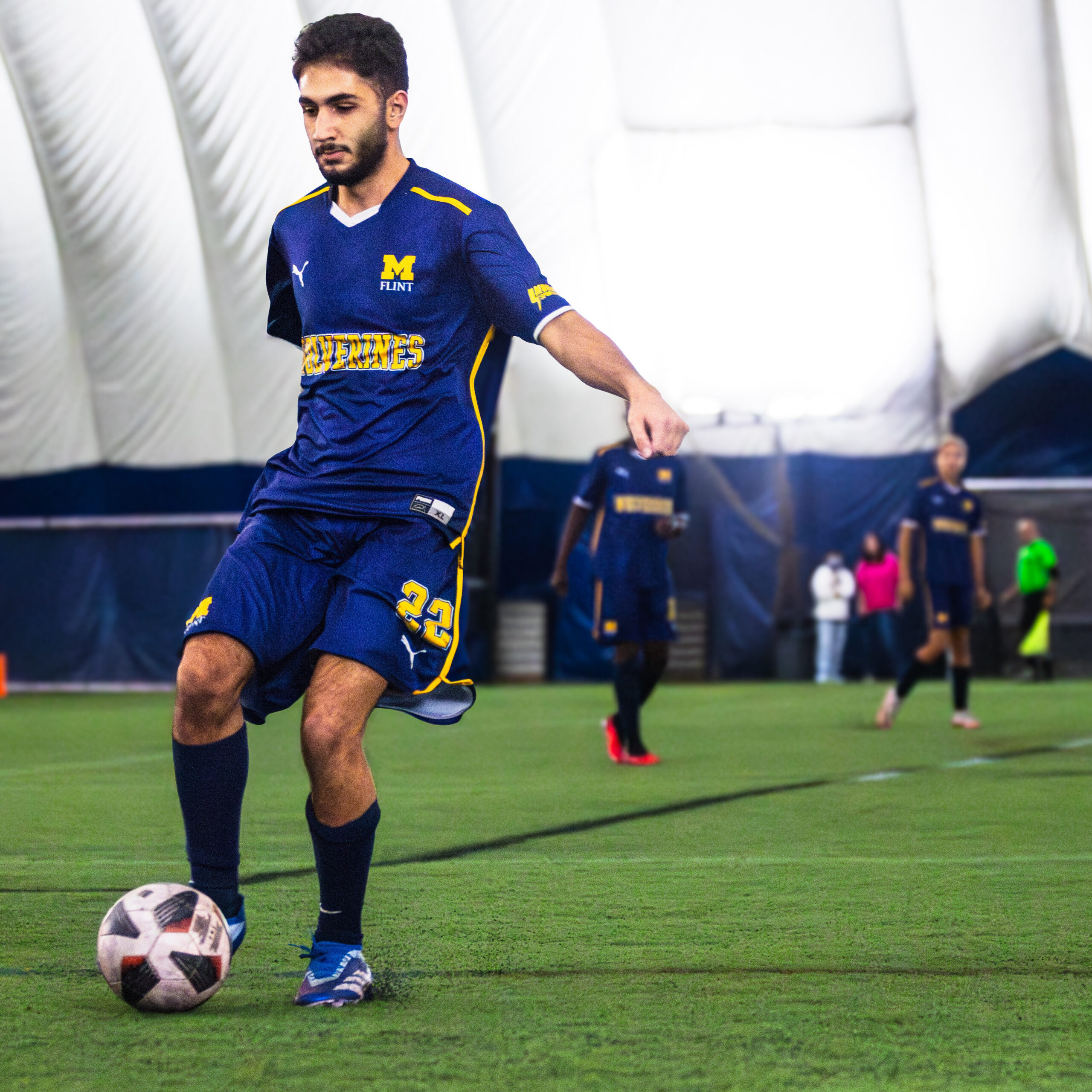 A person in a blue soccer uniform kicks a soccer ball on an indoor field, with other players in the background.