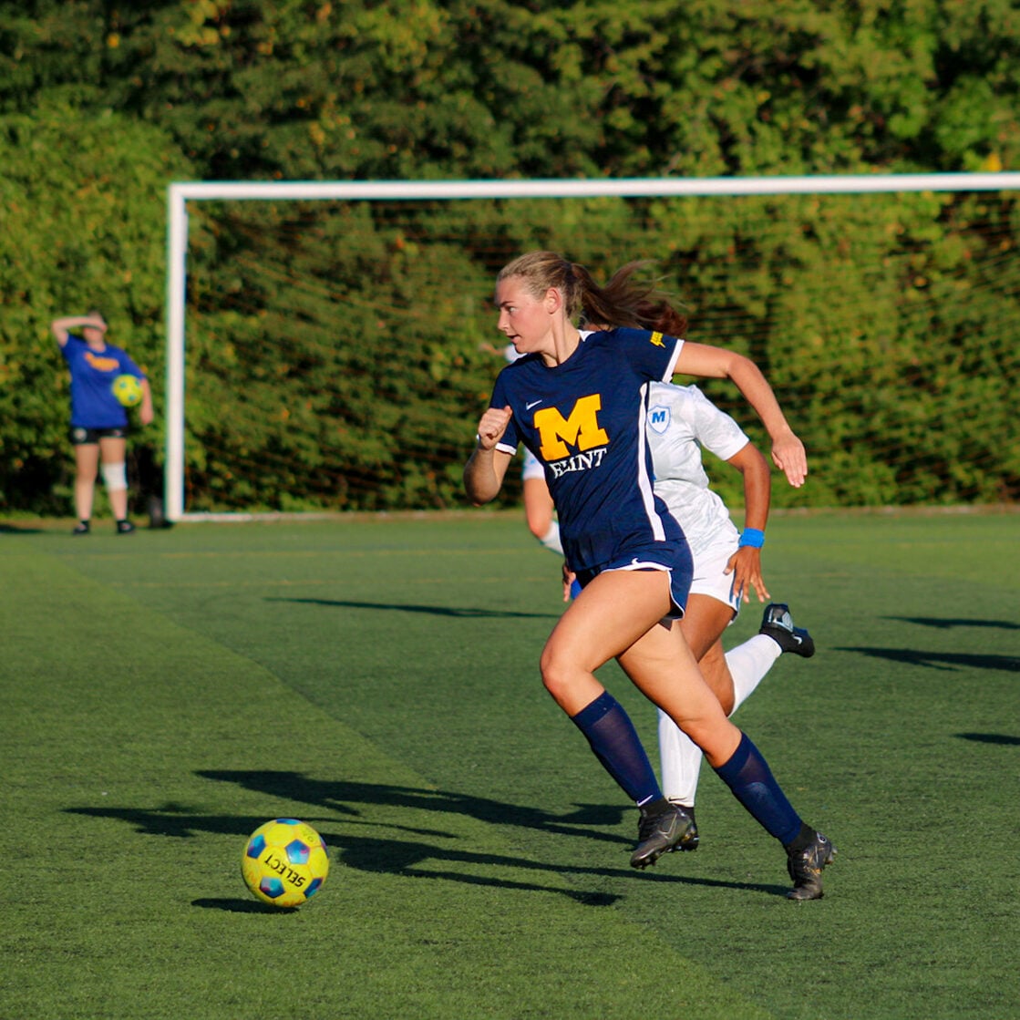 A person in a blue soccer uniform runs with the ball on a field, while another player in white follows closely behind.