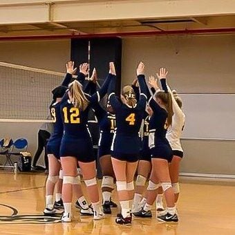 A group of volleyball players huddle together with their hands raised in a gymnasium.