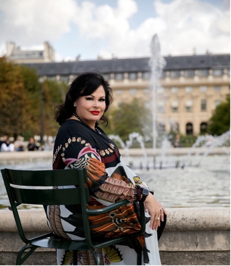 Angeline Boulley sitting on a green chair by a fountain in a park, wearing a colorful shawl and smiling at the camera.
