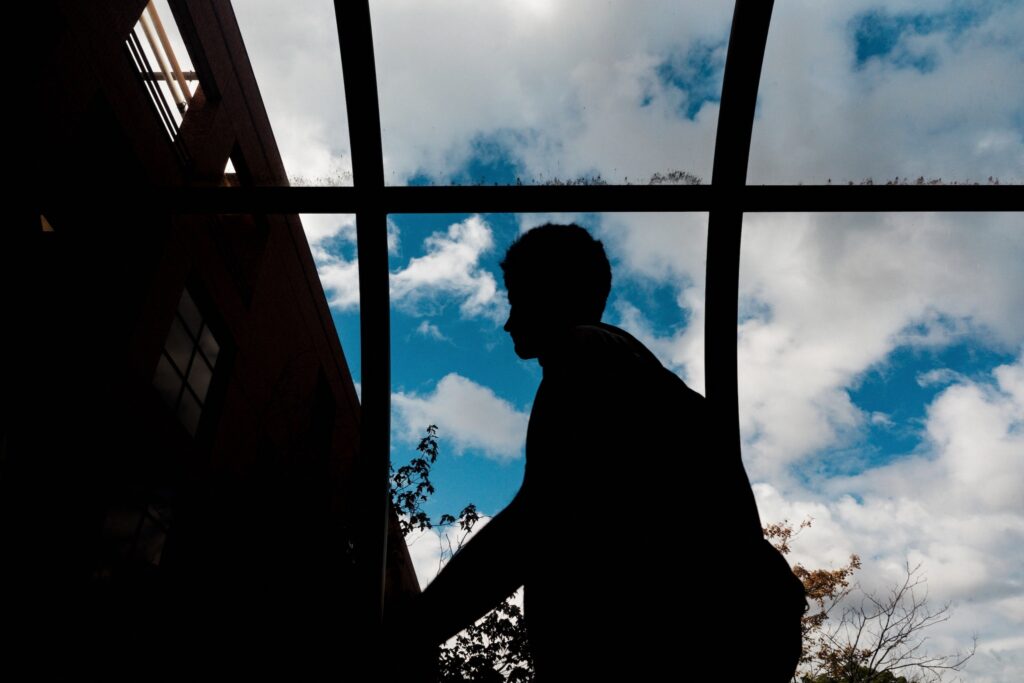A silhouette of a person standing near a window, with blue sky and clouds visible in the background.