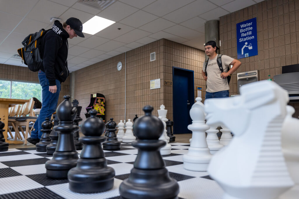 Two people are observing a large outdoor chess set in a common area, with a water bottle refill station visible in the background.