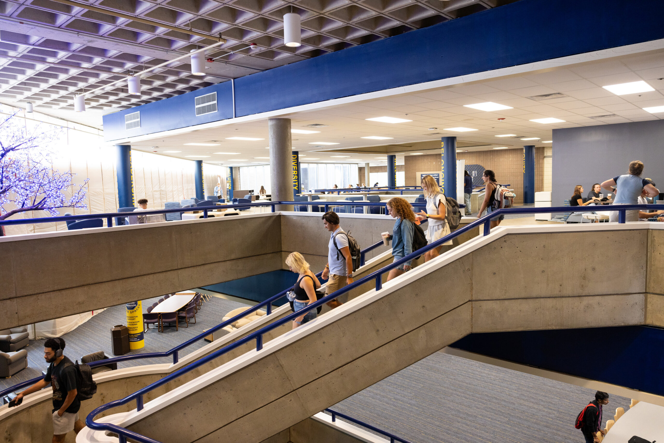 A group of people walking down a staircase in a modern, open space with seating areas and a decorative tree.