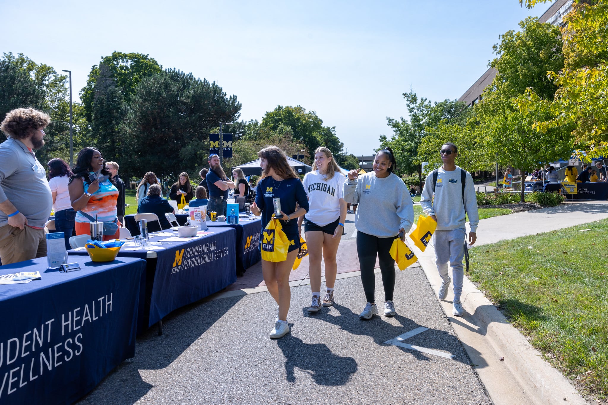 A group of four people walks past tables at an outdoor event, with informational displays about student health and wellness.