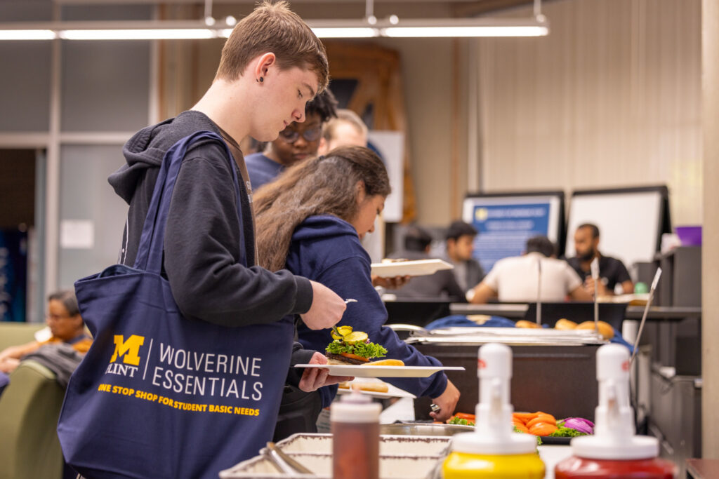 A person with a tote bag is serving food at a buffet, while others are in the background, some seated and others in line.