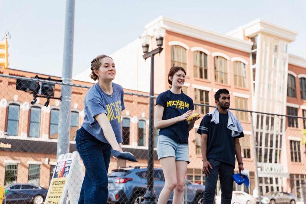 Three people are engaged in an outdoor activity, smiling and interacting, with a cityscape in the background.