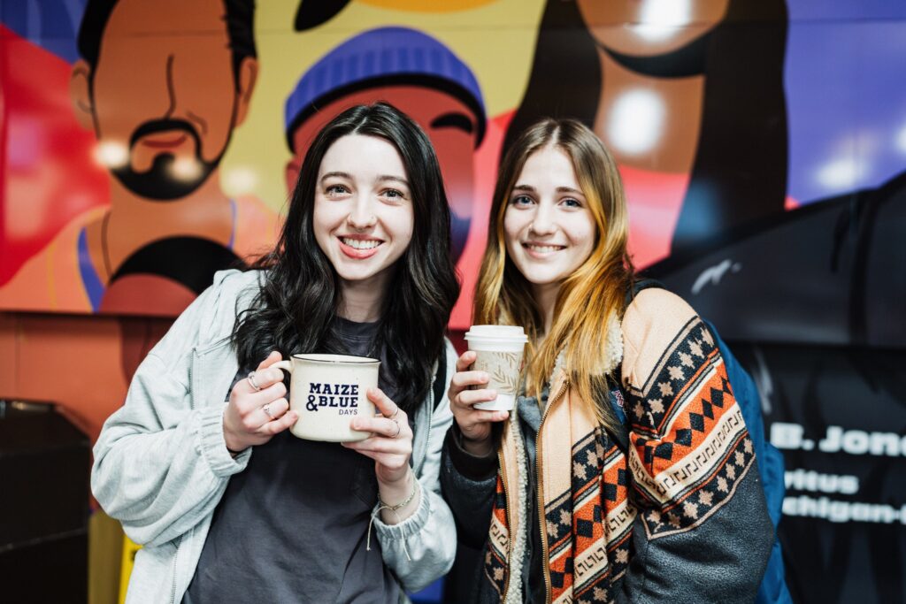 Two people smile while holding coffee cups in front of a colorful mural. One cup has "MAIZE & BLUE" printed on it.