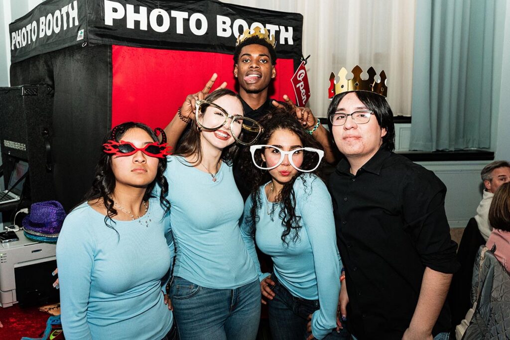 A group of five people poses playfully in front of a photo booth, wearing fun accessories like oversized glasses and crowns.