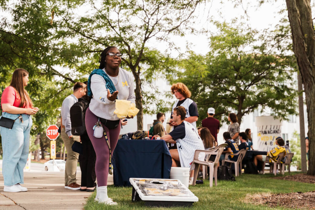 A person throws a bean bag while others socialize at a table during an outdoor event, with trees and a stop sign in the background.