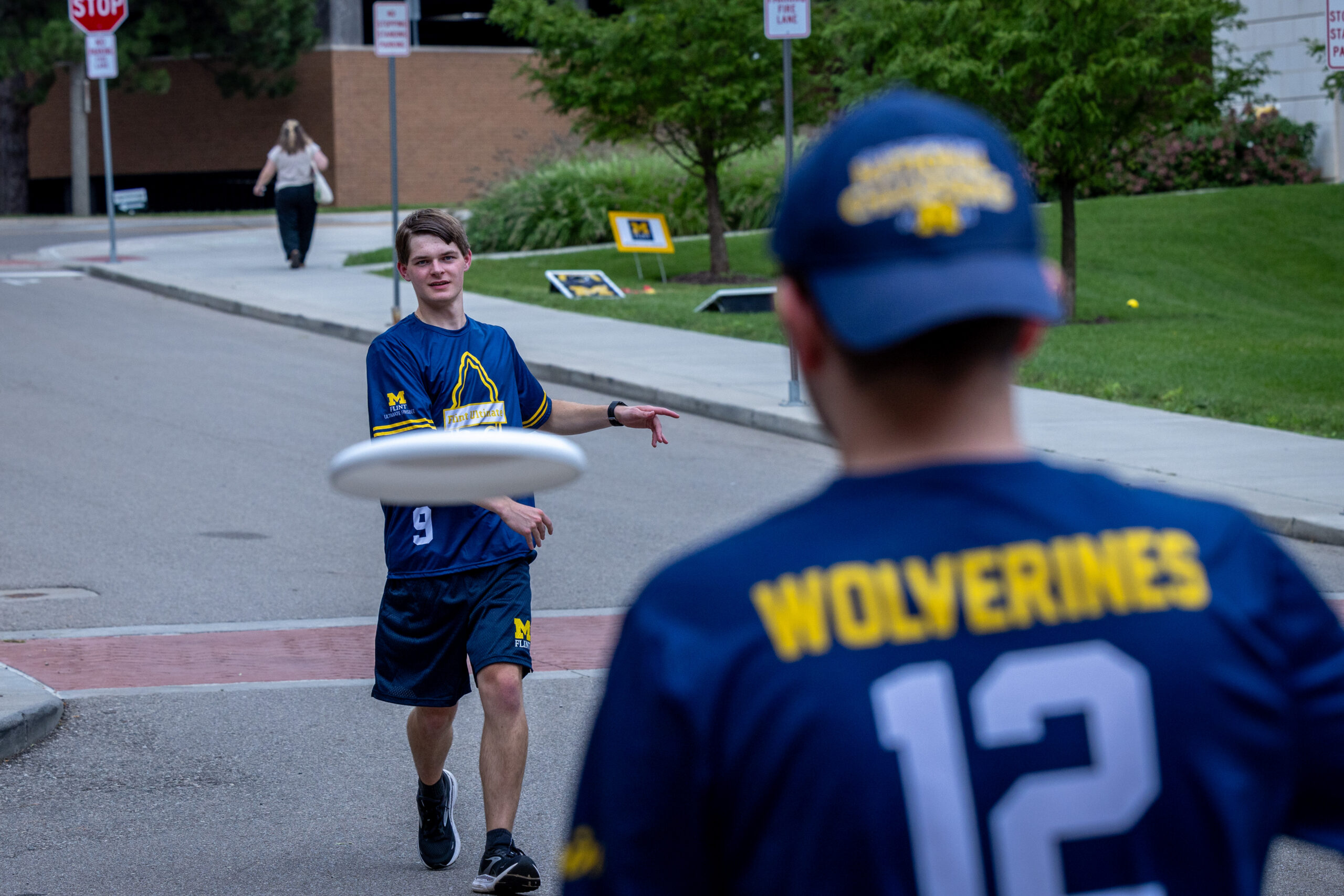 A person throws a frisbee to another person in a sports jersey, with a woman walking in the background on a campus path.
