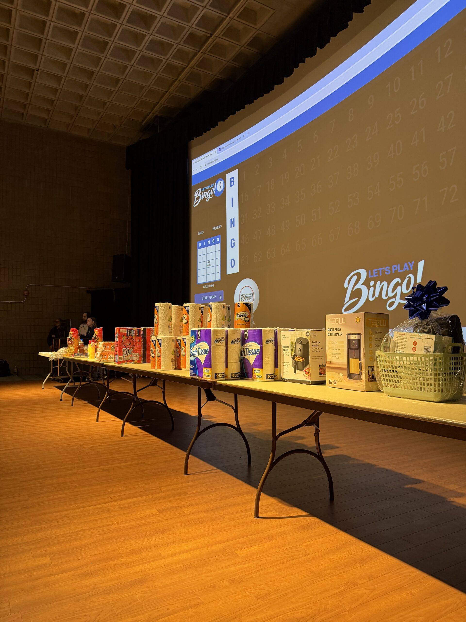 A table displays various boxed items and prizes for a Bingo event, with a Bingo game board projected on a screen behind.