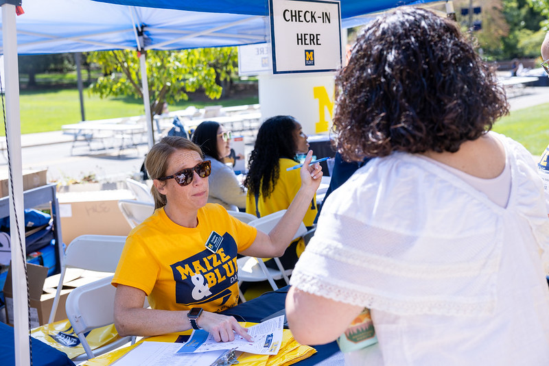 A person at a check-in table wearing a yellow shirt assists another person, with a sign above reading "CHECK-IN HERE."
