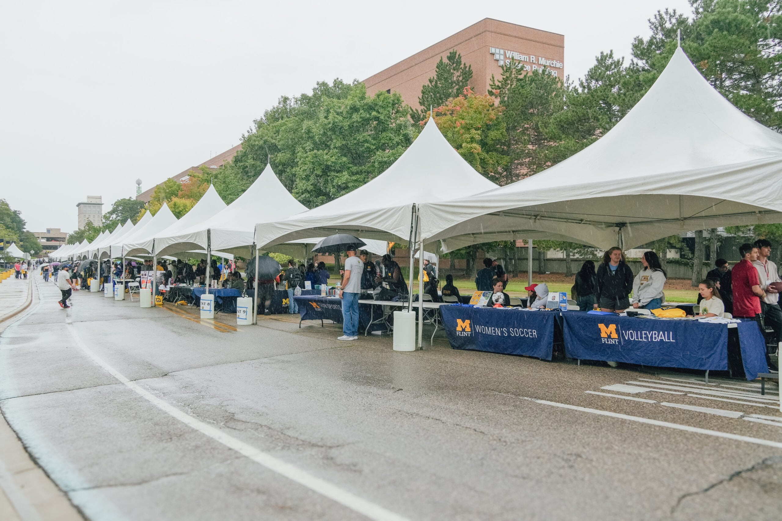 A row of tents at an outdoor event, with tables displaying information for women's soccer and volleyball, under rainy weather.